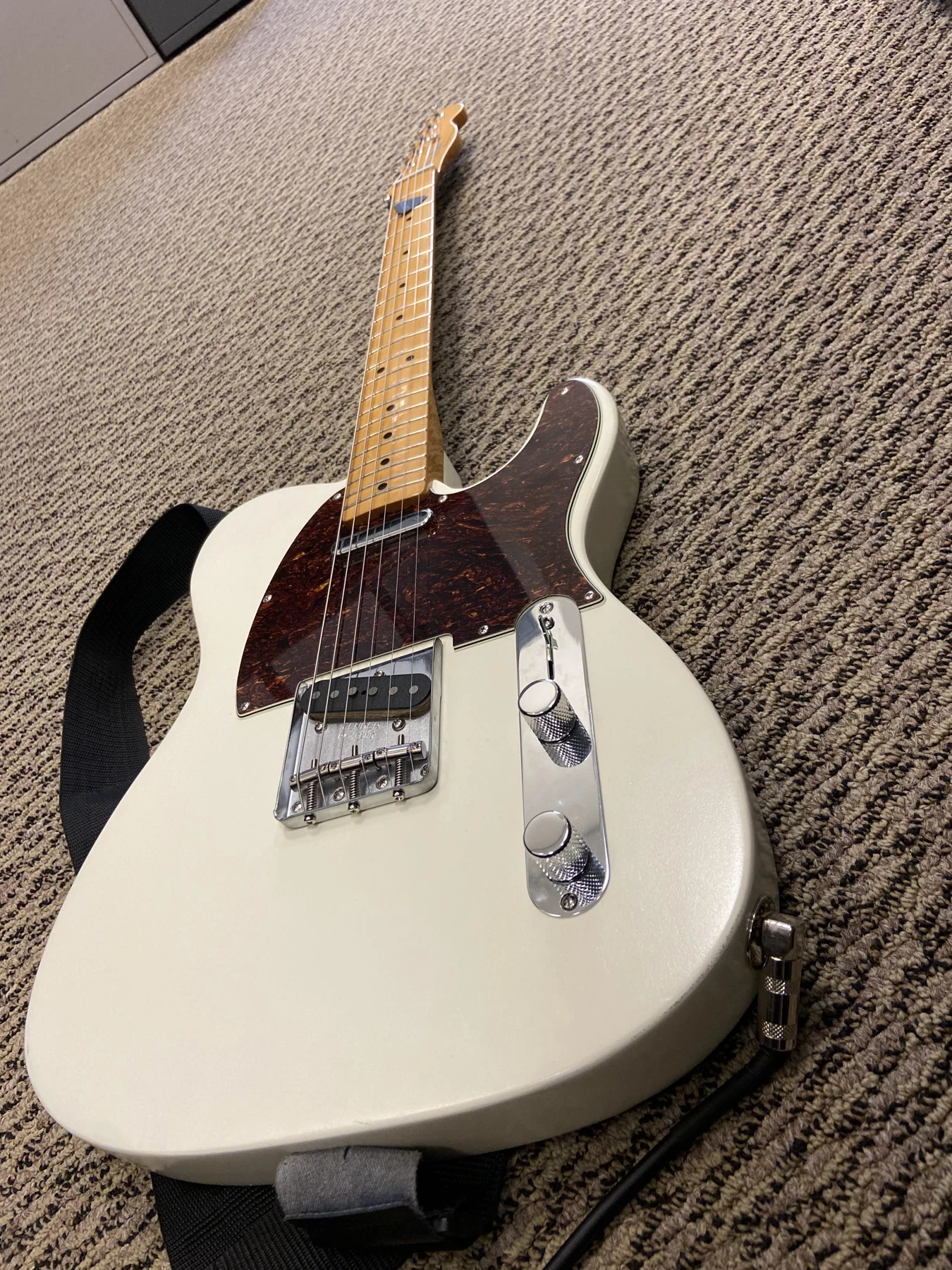 Cream electric guitar with a dark pickguard, maple neck, and chrome hardware, resting on a textured surface